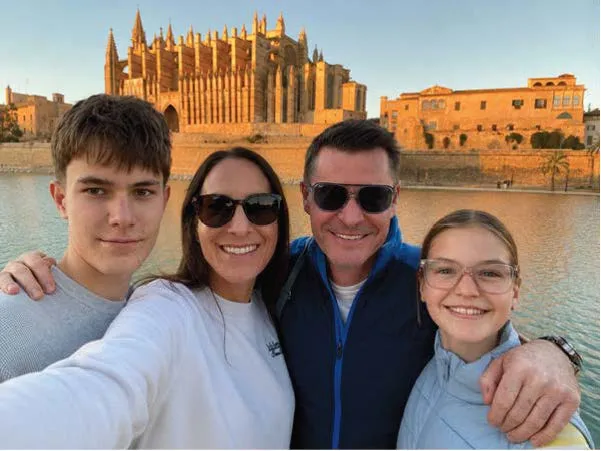 A lady in a white long-sleeved top takes a selfie of her, her husband and her two children while on holiday. Her husband has his arms around his two children and they all smile at the camera. 