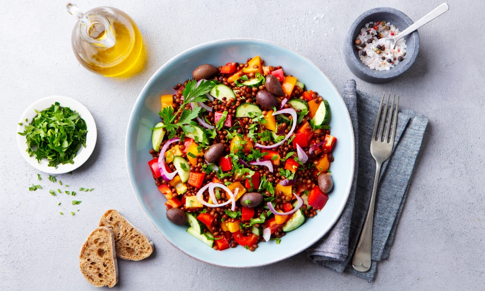 A bowl of lentil salad next to some cutlery, oil, and salad leaves.