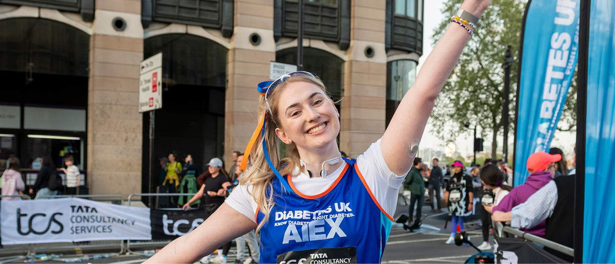 Lady with arms outstretched wearing blue Diabetes UK running vest smiling at the camera. She has blue and orange ribbons in her hair and is wearing a continuous glucose monitor on her arm. 