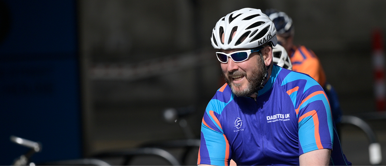Man wearing silver cycling helmet and blue Diabetes UK cycling top riding on a bike.