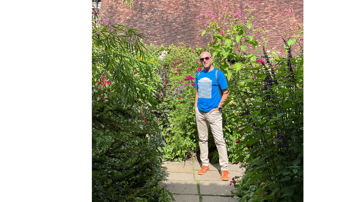 A full body shot of a man wearing a blue T-shirt standing with his hands in his pockets and looking at the camera. He's surrounded by tall green plants. 
