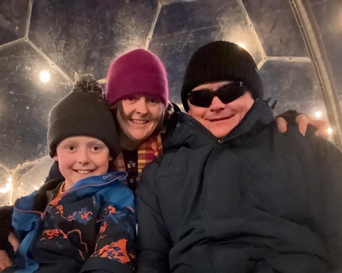 Bruce pictured at a Christmas even with his wife and young son. They are wrapped up in winter wear and there are fairy lights lit in the background.
