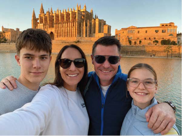 A lady in a white long-sleeved top takes a selfie of her, her husband and her two children while on holiday. Her husband has his arms around his two children and they all smile at the camera. 