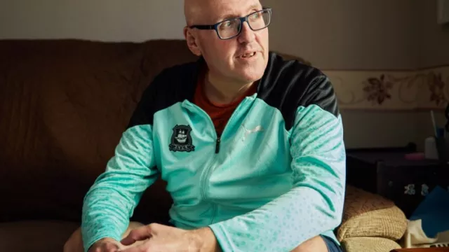 Paul sits in a light blue jacket on a sofa looking to his left