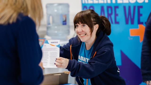 A Diabetes UK employee can be seen handing out a paper resource. The Diabetes UK employee is wearing a pale blue Diabetes UK t-shirt and a navy blue Diabetes UK hoodie. They are smiling at somebody approaching the desk.