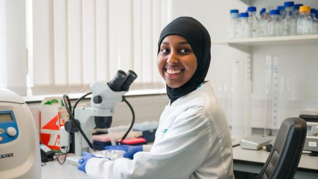 A Black researcher sitting next to a microscope in the lab, smiling