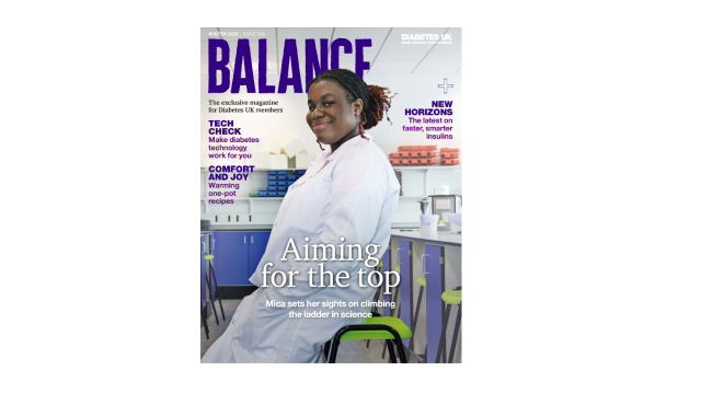 A young woman wearing a lab coat leans against a stool in a research lab smiling at the camera. 