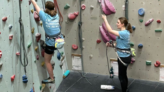 Two people at an indoor climbing wall, one climbing while the other offers support from the ground.