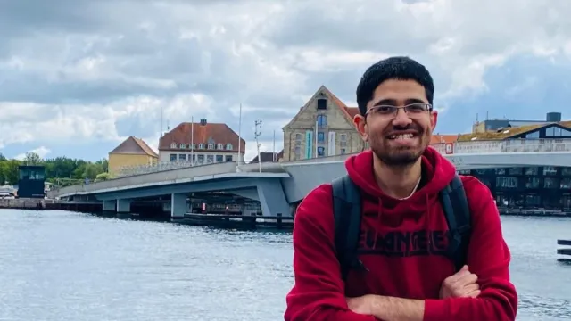 Volunteer Muhammad wears a red jumper, he is standing in front of a river