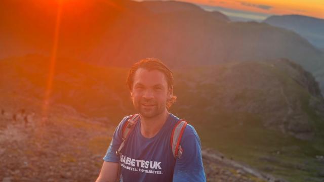 Person wearing a Diabetes UK t-shirt standing on a mountain at sunset, with hills and orange skies in the background.