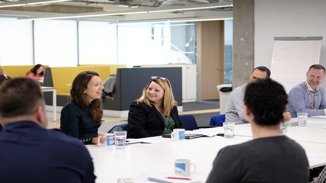 Group of people sat around long white table in office setting smiling and talking