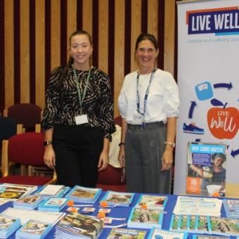 The image shows two ladies next to a banner which reads 'Live Well Hub'. They are standing behind a table with leaflets about diabetes and living well.