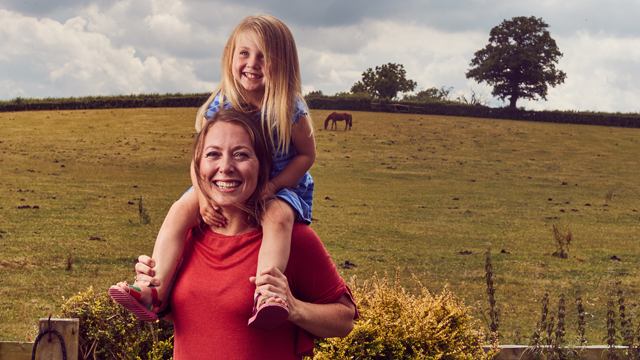 Shannon with her daughter on her shoulders, laughing in the countryside