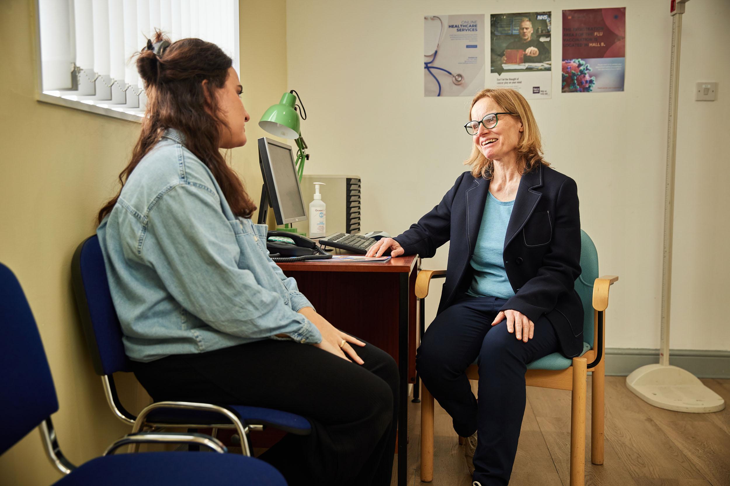 A female doctor is sitting at a desk facing her patient. She has a smile on her face.