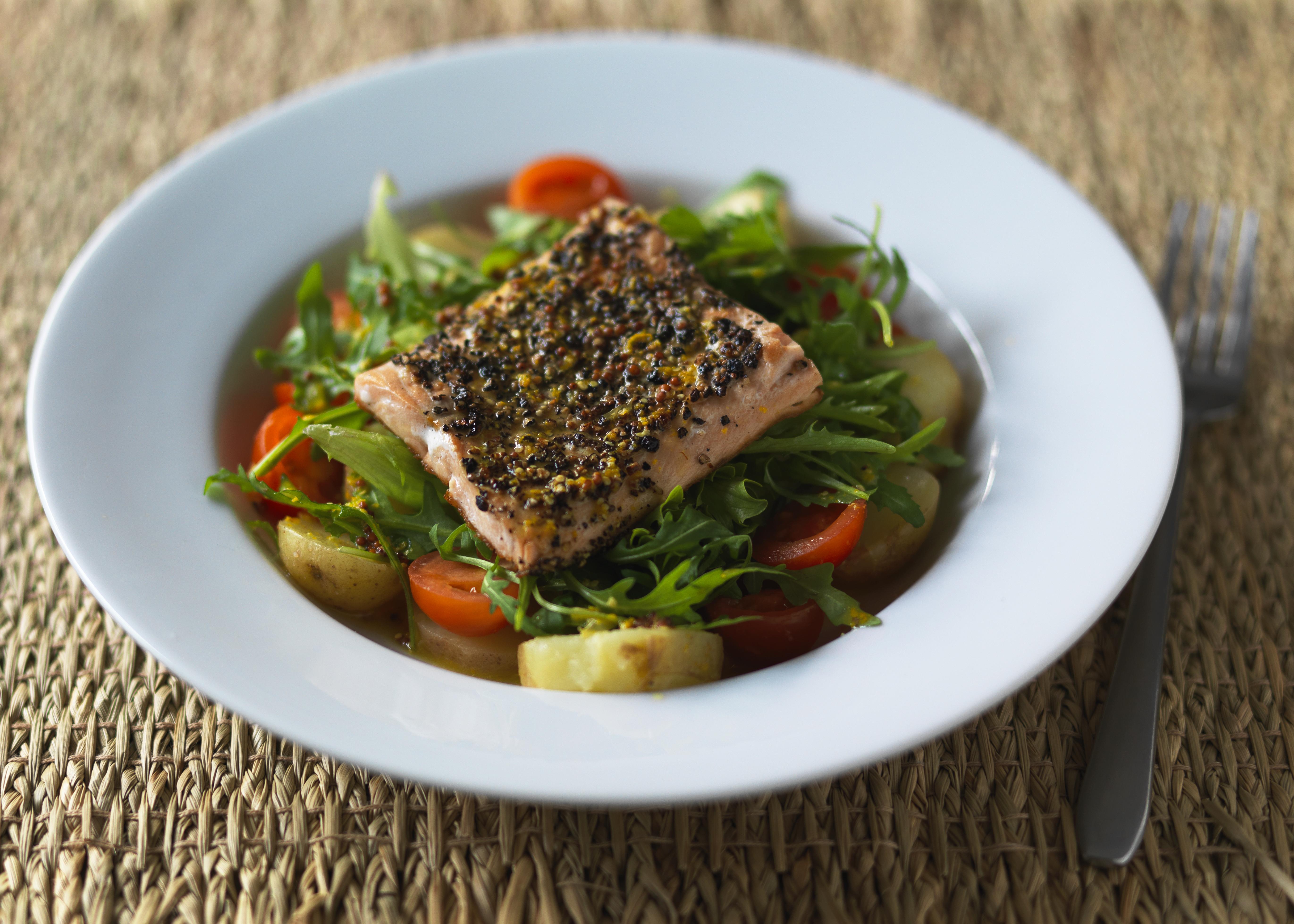 White bowl filled with sliced potatoes, red cherry tomatoes and green salad leaves, topped with a piece of salmon with a crispy skin