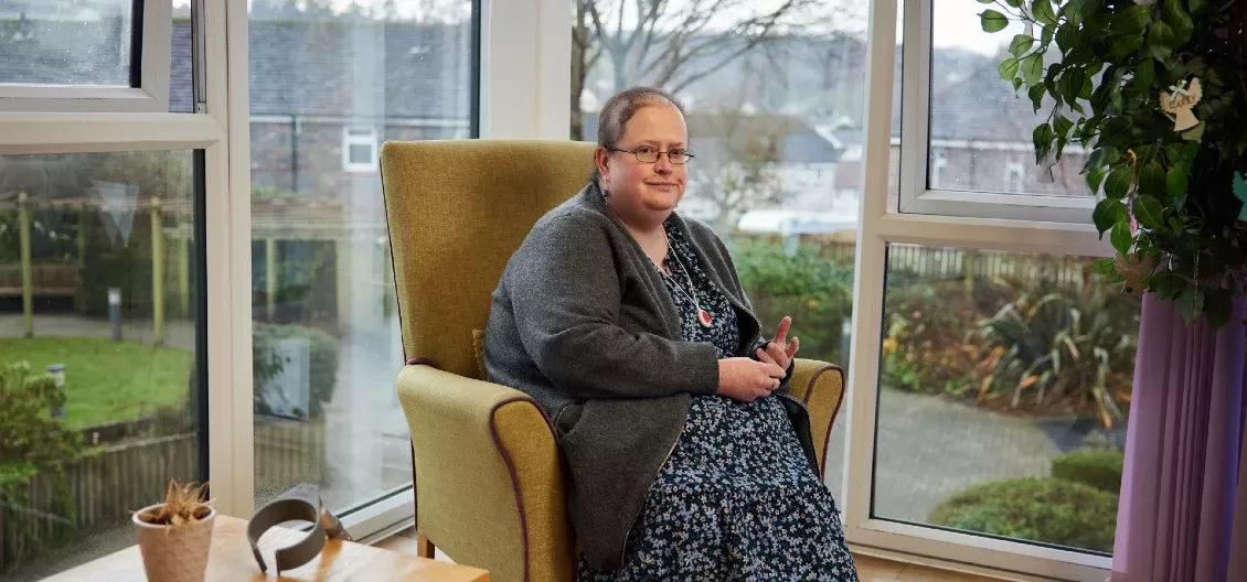 Sarah sits in a yellow armchair in front of big glass windows. She's wearing a dark blue floral dress and a grey cardigan. She's looking into the camera.