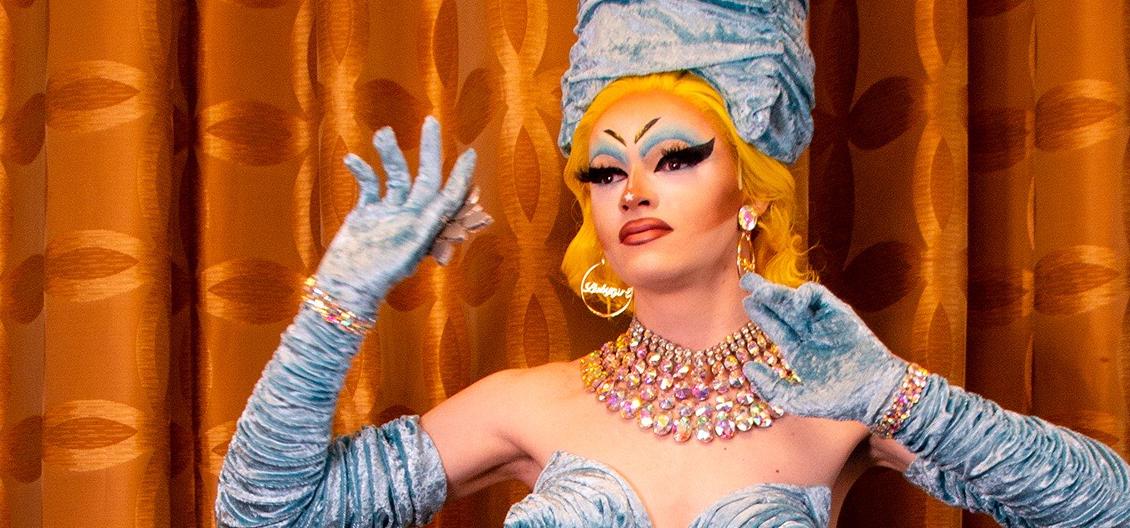 In this image, SALLY is pictured posing towards camera. The picture is framed from head to just below their shoulders. They are a drag artist, with bright blonde hair down to their shoulders. They are wearing pale blue crinkled gloves up to their biceps, with a matching head piece. They are also wearing a jewel necklace, with 6 rows of different jewels. They have hoop gold earrings that read 'babygirl' inside.