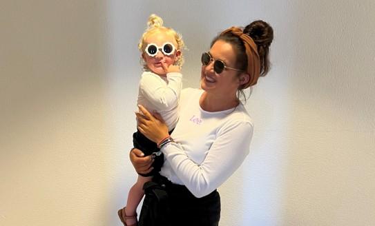 ebony holding her young child, both wearing sunglasses, smiling to camera