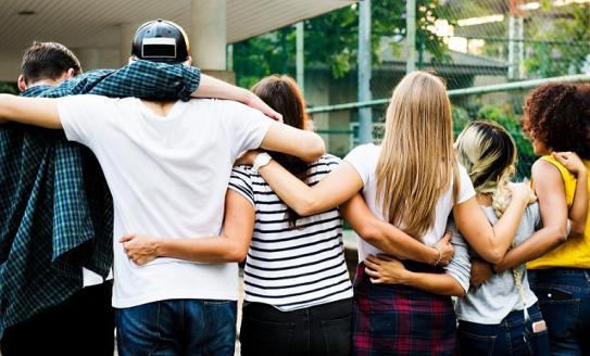 A group of teenagers with their backs to camera, all wearing different outfits and with different hair types, with their arms around eachother