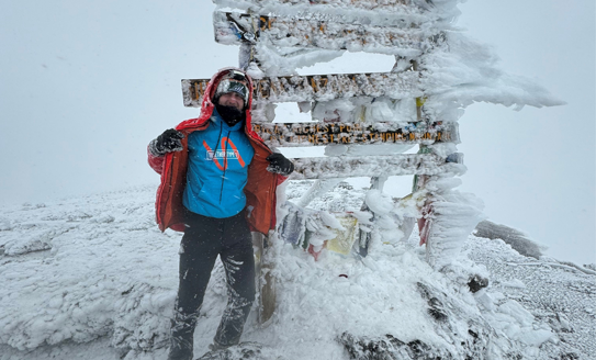 Picture of Michael Cox at the summit of Mount Kilimanjaro. Mike can be seen holding his coat open, revealing a Together Type 1 hoodie. The hoodie is turquoise blue, under an orange coat. Mike is standing in front of a frozen wooden sign where you can see ice frozen sidewards where the wind has blown it. The rocky floor is covered in ice. 