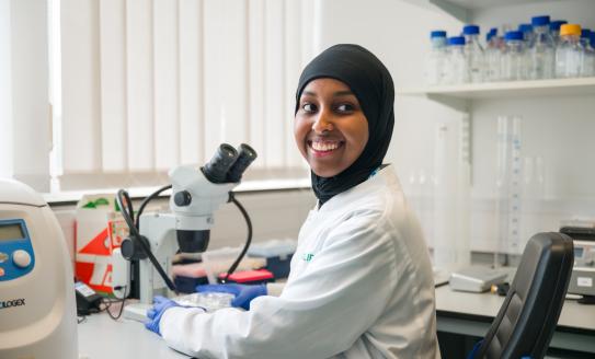 A Black researcher sitting next to a microscope in the lab, smiling
