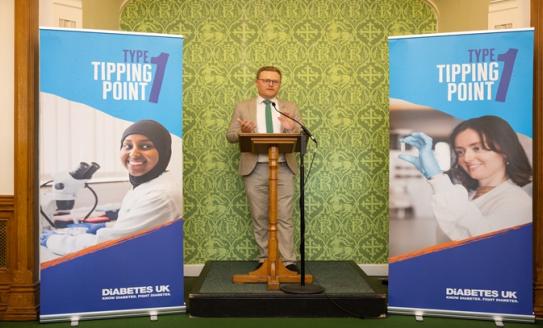 A man in a light brown suit stands before a wooden podium giving a speech. To his left and right, there are banners displaying the 'Type 1 Tipping Point' campaign logo and images of diabetes researchers.