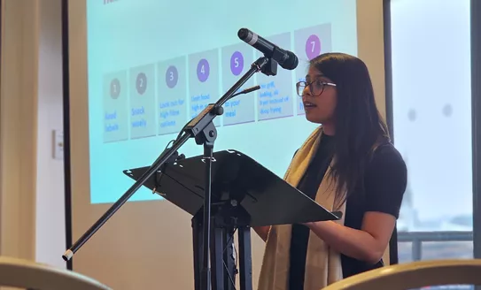 A healthcare professional (female) standing at a lectern and speaking into a microphone. There is a presentation behind her regarding diabetes advice.