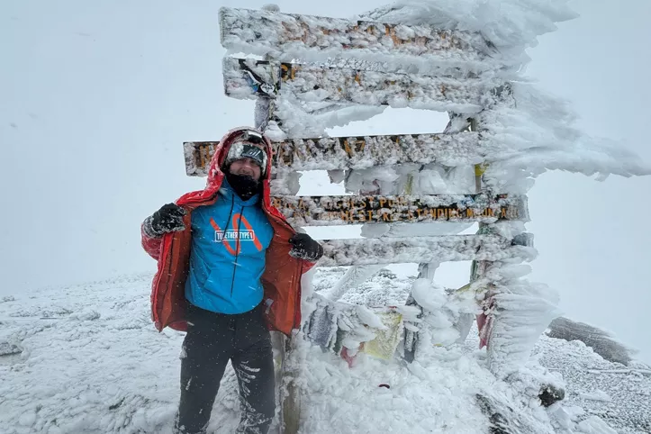 Picture of Michael Cox at the summit of Mount Kilimanjaro. Mike can be seen holding his coat open, revealing a Together Type 1 hoodie. The hoodie is turquoise blue, under an orange coat. Mike is standing in front of a frozen wooden sign where you can see ice frozen sidewards where the wind has blown it. The rocky floor is covered in ice.