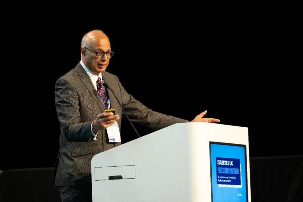 Photo of a man dressed in a grey suit gesturing and speaking on a podium