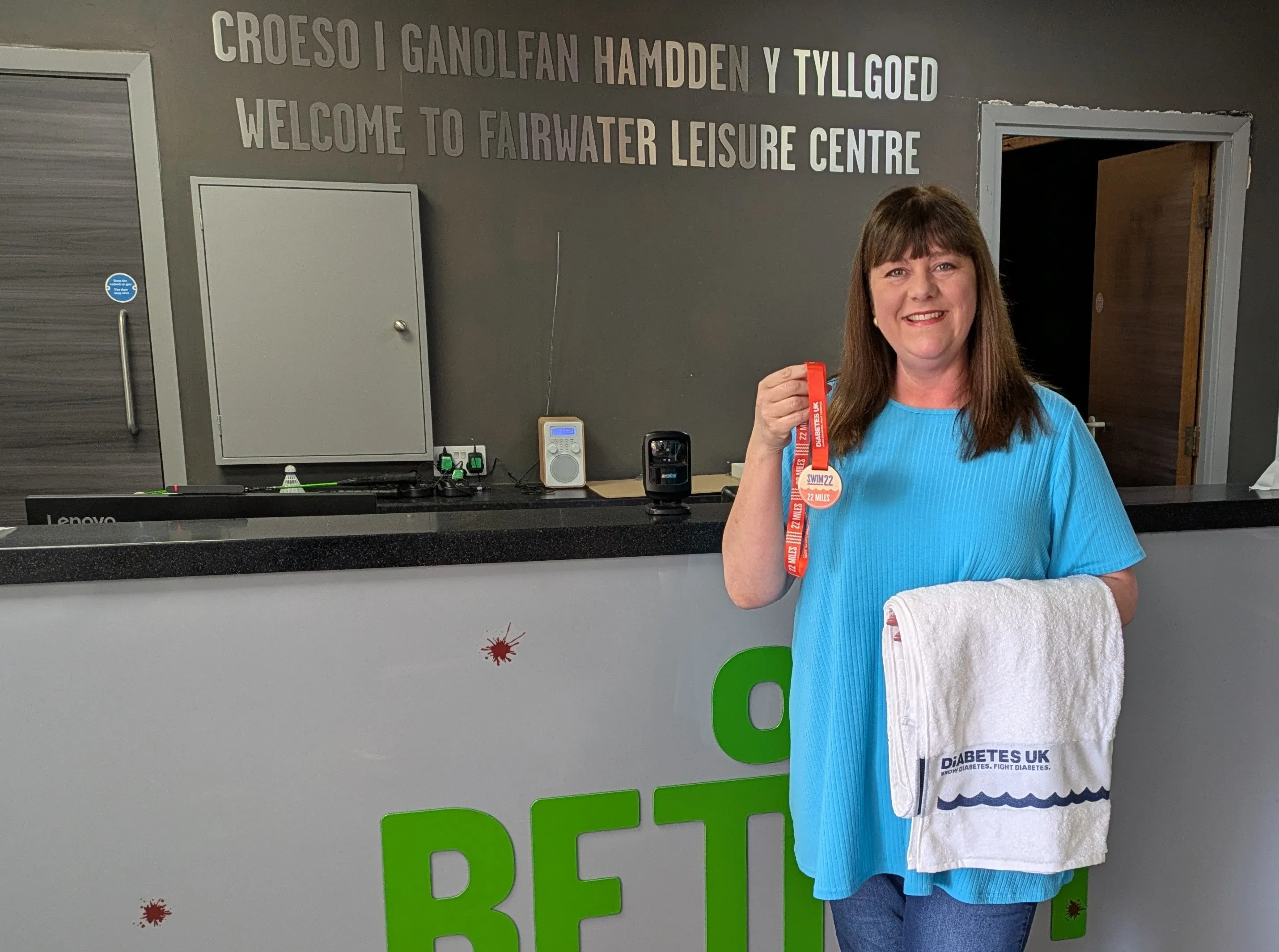 Swim22 participant Julie standing in front of reception desk holding towel and medal 
