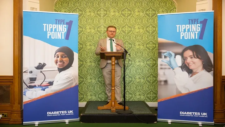 A man in a light brown suit stands before a wooden podium giving a speech. To his left and right, there are banners displaying the 'Type 1 Tipping Point' campaign logo and images of diabetes researchers.