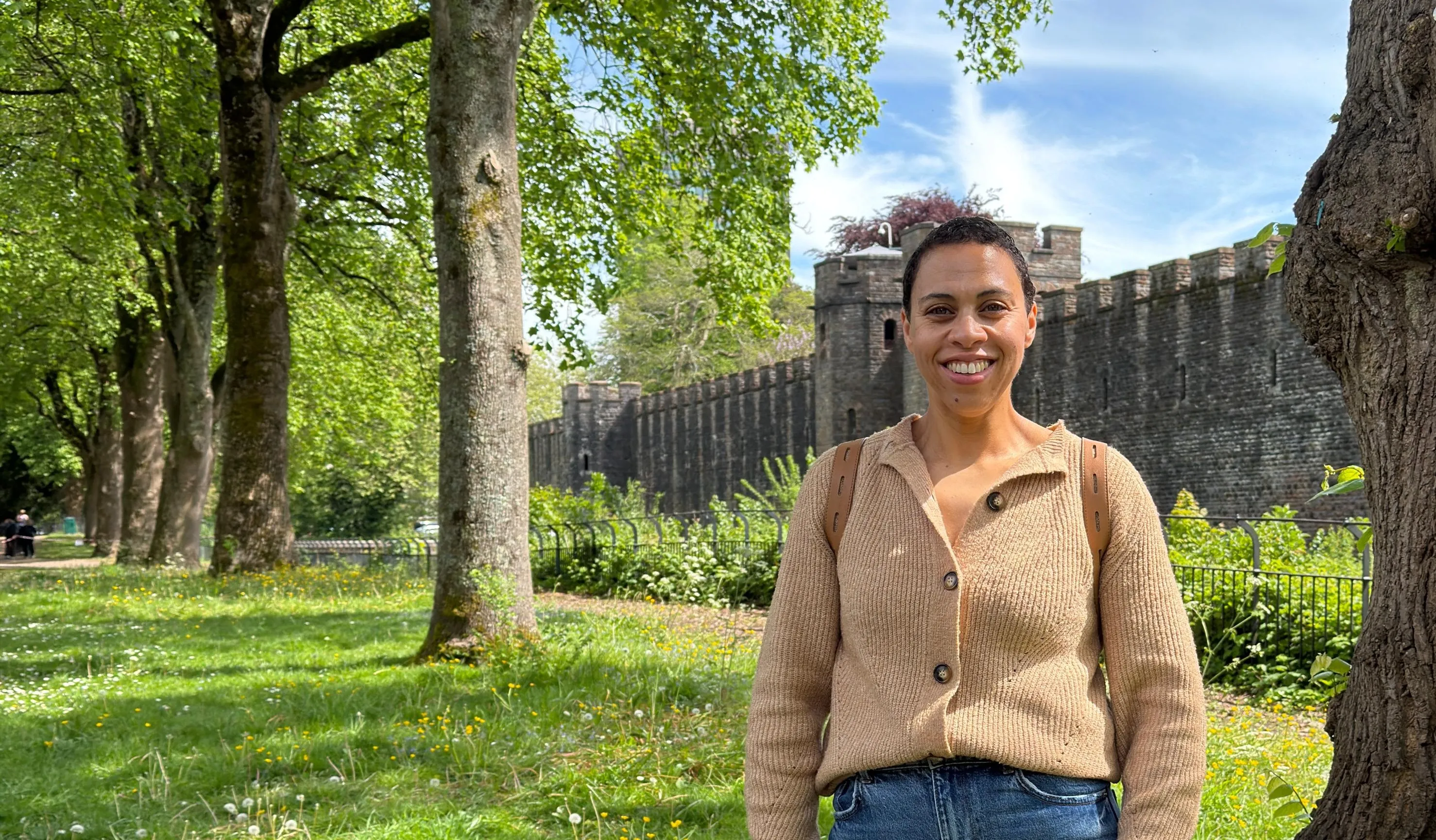 A lady with short black hair wearing a beige sweater, pictured in a park on a sunny day