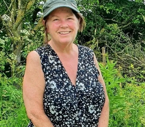 A lady pictured in a short sleeved black top and grey hat with lots of greenery in the background. 