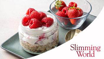 A glass jar with a wheat biscuit base and yoghurt and raspberries on top. Next to the jar is a small glass bowl with strawberries and raspberries. Both are placed on a grey rectangular plate.