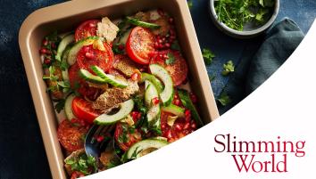 Halved tomatoes, cucumber slices, pomegranate seeds, and bite-sized pitta pieces in a baking tray with a dark blue background. A bowl of fresh chopped parsley is on the side.