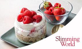 A glass jar with a wheat biscuit base and yoghurt and raspberries on top. Next to the jar is a small glass bowl with strawberries and raspberries. Both are placed on a grey rectangular plate.