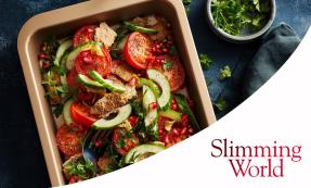 Halved tomatoes, cucumber slices, pomegranate seeds, and bite-sized pitta pieces in a baking tray with a dark blue background. A bowl of fresh chopped parsley is on the side.