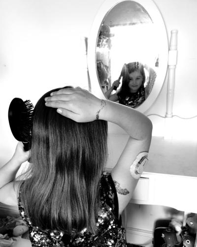 Ava, who lives with type 1 diabetes, brushing her hair in the mirror with her back to camera. Her continuous glucose monitor is visible on the back of her arm.