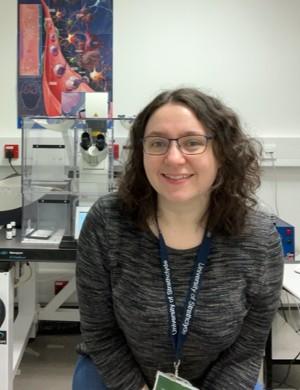 Holly sitting in front of a microscope in her lab. She is smiling to camera, wearing a grey top and a lanyard