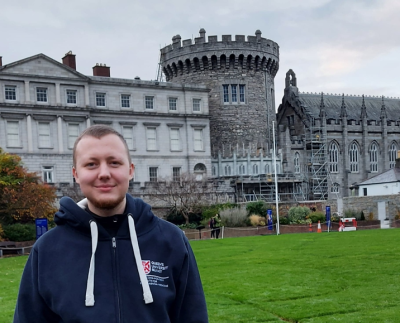 Adam wearing a navy blue hoody, standing in front of a grey castle on some grass, smiling to camera