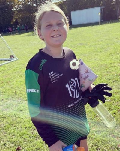 A girl called Skye, who plays football as a Goalkeeper living with type 1 diabetes. She is in a green and black goalkeepers football kit, standing up on the grass, holding her gloves, water and drinks bottle whilst smiling