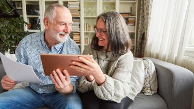 An image of two older individuals sitting on a couch, holding a tablet device together that they are both looking at.